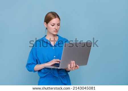 Serious beautiful young woman in casual blue shirt dress posing standing, working on laptop, doing work, isolated on blue background, studio portrait. Caucasian girl freelancer