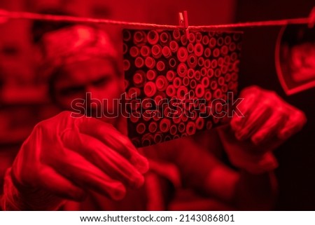 Close-up shot of young photographer wearing protective gloves hanging wet developed photos on thread in red lit darkroom