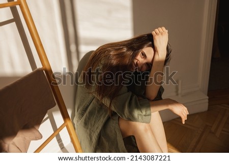 Cute caucasian young woman covers her face with hand from sun while sitting in light room. Brunette in casual home clothes looks at camera. Happy weekend concept Royalty-Free Stock Photo #2143087221
