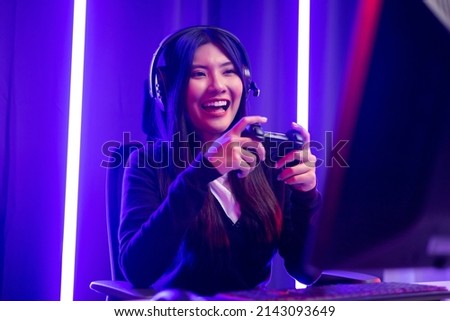 Playing video game. Young asian pretty woman sitting on chair holding joystick in living room. Happy female Professional Streamer chinese wearing headphone playing game in dark room neon light.