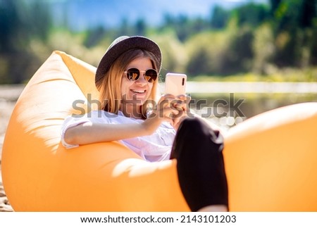 Portrait of charming smiling woman in sunglasses using cellphone and write messages to friends while lying on inflatable mattress on summer beach Royalty-Free Stock Photo #2143101203