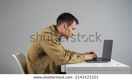 Young man works on laptop sitting at white table in office against grey wall. Focused employee in mustard jacket with crooked neck side view Royalty-Free Stock Photo #2143121521
