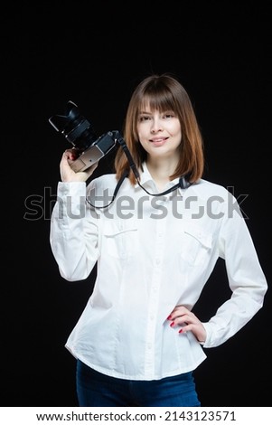 Portrait of a young woman wearing a white shirt holding a camera in her hand. The concept of a successful photographer. Isolated black background.