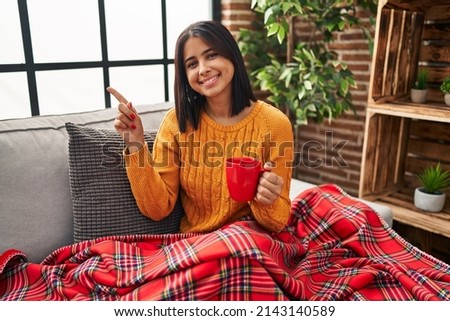 Young hispanic woman sitting on the sofa drinking a coffee at home with a big smile on face, pointing with hand finger to the side looking at the camera. 