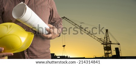 torso worker or engineer hand holding yellow helmet and blueprint on background of new highrise apartment buildings and construction cranes on background of sunset sky Silhouette Crane lifts load