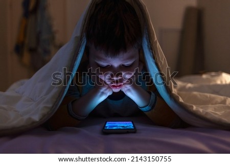 Little boy on cell phone under blanket. Curious boy sits in bed under white blanket and play games on smartphone in the dark room.  Image of nice boy in pajama sitting in front of mobile.  Royalty-Free Stock Photo #2143150755