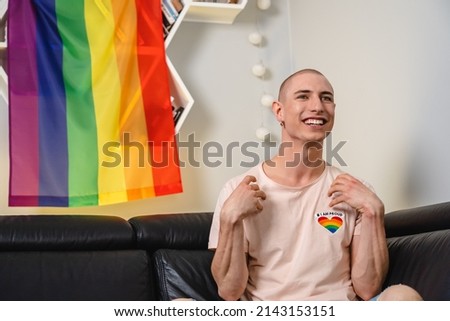 Bald queer caucasian activist sitting on a sofa, watching tv, and smiling with their hands up. Rainbow flag in the background. High quality photo