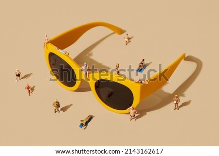 some miniature people, wearing swimsuit, relaxing on top of a pair of yellow plastic-rimmed sunglasses and some more miniature people standing around, against a pale brown background Royalty-Free Stock Photo #2143162617