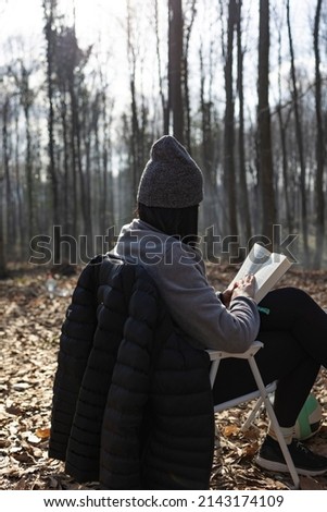 Young girl with hat reading a book in forest. Back view. Unrecognizable person.	 Vertical photo.