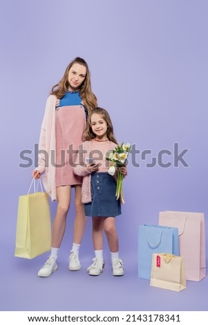 full length of kid holding flowers and using smartphone while standing with mother near shopping bags on purple