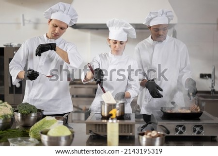 Multiracial team of three cooks in uniform cooking together in the professional kitchen. Latin cook frying meat and european guys making sauce. Concept of teamwork at restaurant Royalty-Free Stock Photo #2143195319