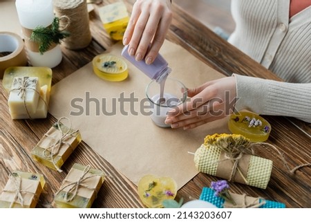 Top view of craftswoman pouring liquid wax in glass near soap bars and craft paper Royalty-Free Stock Photo #2143203607