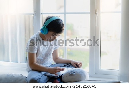 Happy young boy wearing headphone for playing game online on internet with friends, Kid sitting next to window reading or watching cartoon on tablet Child relaxing at home in the morning on Springtime