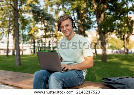 Excited caucasian guy relaxing with laptop and headphones in park, sitting on bench and smiling at camera, spending time outdoors, studying and listening music