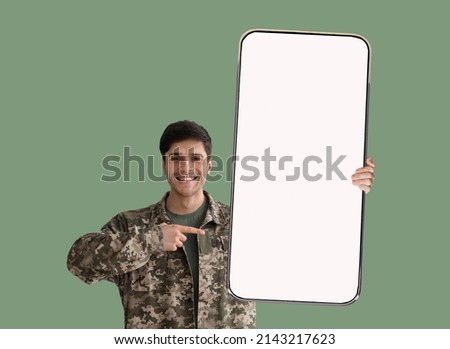 Handsome young military guy in uniform showing nice brand new smartphone with empty screen, pointing at white display and smiling, recommending mobile app, green studio background, mockup, copy space
