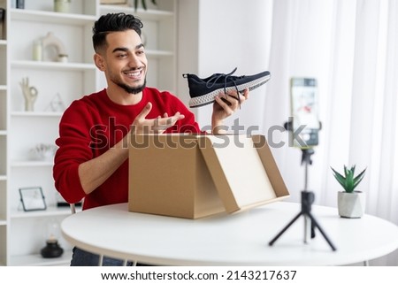 Fashion Blog. Young Arab Male Influencer Recording Video Content At Home, Smiling Middle Eastern Guy Unpacking Box With New Shoes In Front Of Camera, Using Smartphone On Tripod, Copy Space