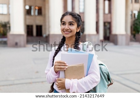 Outdoor portrait of cheerful indian female student with backpack and workbooks standing near college building, looking at camera and smiling