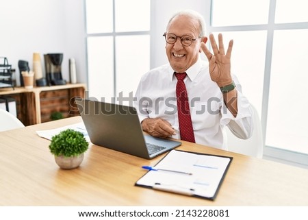 Senior man working at the office using computer laptop showing and pointing up with fingers number four while smiling confident and happy. 