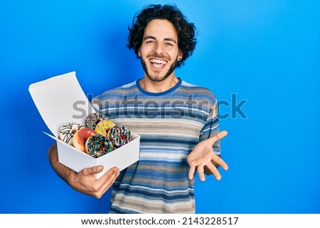 Handsome hispanic man holding tasty colorful doughnuts celebrating achievement with happy smile and winner expression with raised hand 