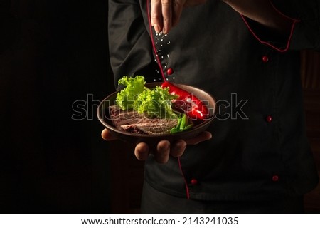 A professional chef sprinkles salt on a sliced beef steak and vegetables in a plate. The concept of serving dishes to order.