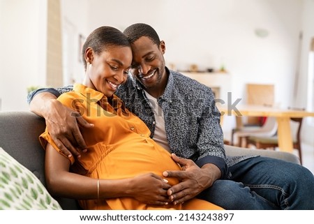 Happy african american husband and pregnant woman hugging belly. Smiling black man hugging happy pregnant wife sitting on sofa and holding tummy. Mature loving couple expecting their first baby. Royalty-Free Stock Photo #2143243897