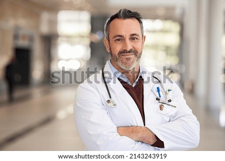 Portrait of happy doctor standing with folded hands in modern hospital corridor. Successful general practitioner looking at camera with satisfaction at private clinic. Confident proud head physician. Royalty-Free Stock Photo #2143243919