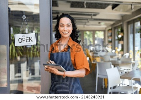 Portrait of happy waitress standing at restaurant entrance and holding digital tablet. Happy young latin woman owner in apron standing at coffee shop entrance, leaning while waiting clients. 