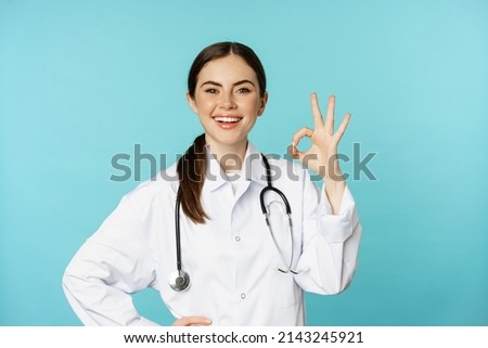 Portrait of satisfied, smiling medical worker, woman doctor showing okay, ok, zero no problem gesture, excellent sign, standing pleased over torquoise background