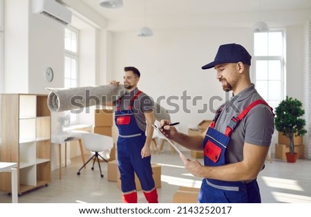 Two workers in uniforms remove things from old apartment. Happy man holding clipboard and taking notes while his teammate is carrying rugs and other stuff. Moving house, using delivery service concept Royalty-Free Stock Photo #2143252017