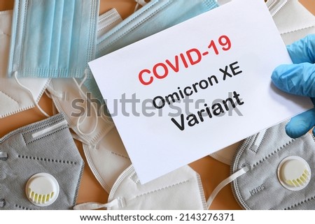 Doctor's hand in blue glove with white paper and text Covid-19 Omicron XE Variant with various protection masks on background. COVID-19 Omicron XE Variant strain protection concept. Royalty-Free Stock Photo #2143276371