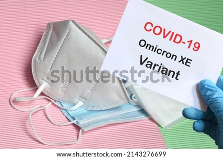 Doctor's hand in blue glove with white paper and text Covid-19 Omicron XE Variant with various protection masks on background. COVID-19 Omicron XE Variant strain protection concept. Royalty-Free Stock Photo #2143276699