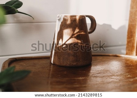 Home aesthetics. Modern ceramic kettle in sunlight on rustic wooden chair and leaves shadow. Stylish ceramic minimalist vase in sunny boho room with plants.