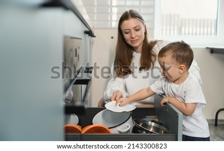 Happy mom and cheerful baby in the kitchen. A restless child sorts through the dishes in the kitchen drawer. Mother's love. Motherhood.