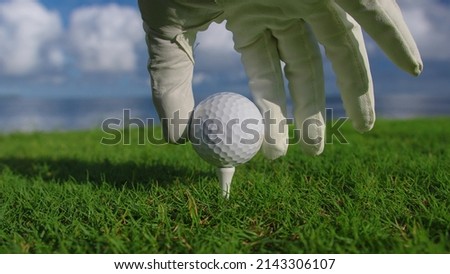 Male hand in a golf glove puts the ball on the field on the background of the blue ocean. 