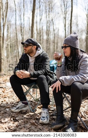 Young man and woman sitting on chairs in forest. They are drinking hot tea. Blur trees background. Vertical photo.