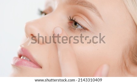 Extreme close-up of young Caucasian woman with pure skin applies face serum smearing drops on her cheek on white background | Skin care serum commercial concept Royalty-Free Stock Photo #2143310409