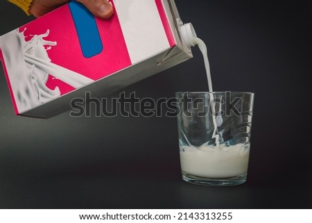 A person's hand holding a carton of delicious fresh milk and pouring it into a glass. Image of a stream of milk filling a small glass on a black background. Royalty-Free Stock Photo #2143313255