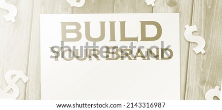 Build Your Own Brand words on a page and paper dollar signs around on wooden table. Branding rebranding marketing business concept.