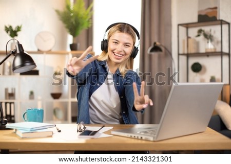 Joyful pretty young blond woman wearing headphones, sitting at the table at modern apartment, and showing victory sign gesture to camera. Smiling girl working on laptop at home
