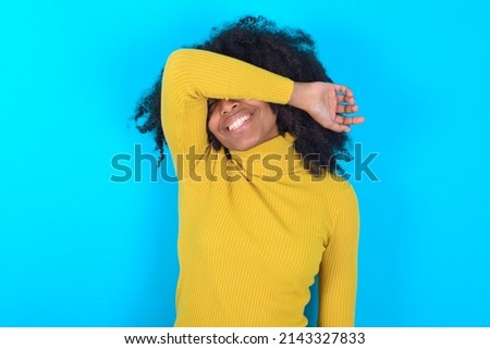 Young woman with afro hairstyle wearing yellow turtleneck over blue background covering eyes with arm smiling cheerful and funny. Blind concept.