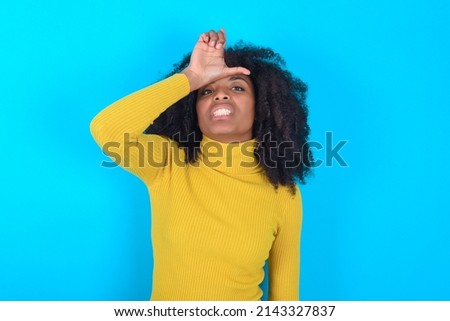 Young woman with afro hairstyle wearing yellow turtleneck over blue background making fun of people with fingers on forehead doing loser gesture mocking and insulting.