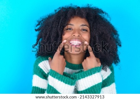 Happy Young woman with afro hairstyle wearing striped sweater over blue background with toothy smile, keeps index fingers near mouth, fingers pointing and forcing cheerful smile