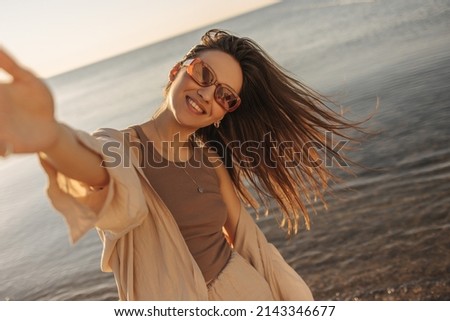 Young fair-skinned happy lady makes selfie in sunglasses on background of sea. Brown-haired woman is waving her hair in upbeat cheerful mood. Leisure, relaxation, activity and lifestyle concept.