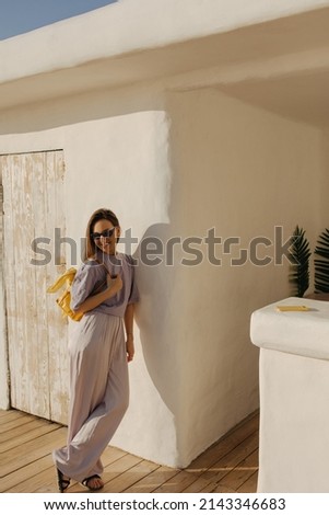 In full growth, slender young european lady stands cross-legged, leaning against white wall. Smiling brown-haired woman in sunglasses is dressed loose summer clothes with bag.