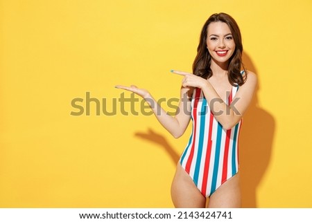 Happy young woman wear red blue swimsuit pointing fingers hands aside on copy space mock up promo area isolated on vivid yellow color wall background studio. Summer hotel pool sea rest sun tan concept