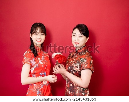 Portrait of asian young daughter and mother wearing traditional cheongsam qipao and smiling with flowers carnation in hands isolated on red background.