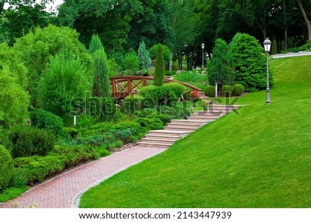 iron ground lantern and poli garden lighting of park with stone stairs and path paved tiles with drain lattice in park among plants, evergreen bushes and trees on slope hill green lawn, nobody. Royalty-Free Stock Photo #2143447939