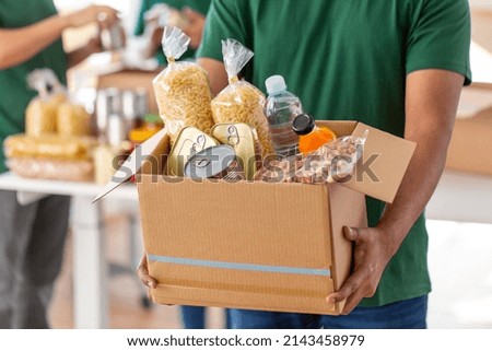 charity, donation and volunteering concept - close up of male volunteer's hands holding box with food over group of people at distribution or refugee assistance center Royalty-Free Stock Photo #2143458979
