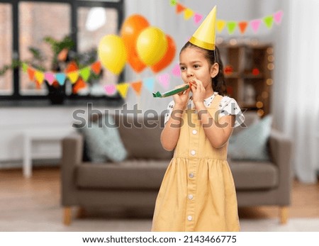 birthday, childhood and people concept - portrait of smiling little girl in party hat with blower having fun over decorated home room background