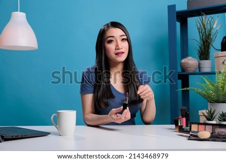 Asian content creator holding lipstick talking with remote subscribes while recording makeup tutorial for vlogging channel in studio with blue background. Vlogger advertising product